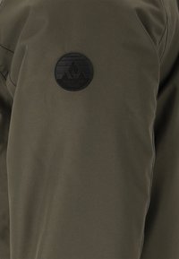 Olivgrüne Jacke aus strukturiertem Material mit runder schwarzer Logopatch mit Bergdesign und gestickten Details auf der Schulter.
