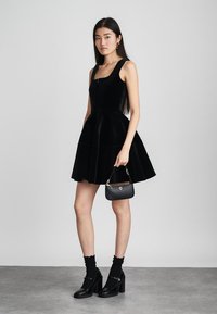 Vestido de terciopelo negro con un corsé ajustado y falda acampanada, combinado con un pequeño bolso negro, medias hasta la rodilla y zapatos de tacón robusto.