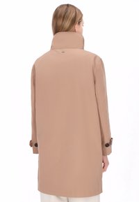 Beige vandafvisende frakke med høj krave, lange ærmer og manchetter med knapper. Glat tekstur og minimalistisk design uden lommer.