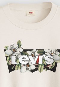 Felpa color crema con una grafica floreale caratterizzata da fiori di magnolia bianchi e foglie verdi, accanto al logo "Levi's" in lettere in grassetto.