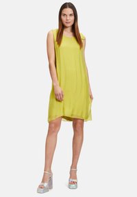 Limettengrünes ärmelloses Kleid mit einem fließenden Design, rundem Ausschnitt und leicht asymmetrischem Saum. Kombiniert mit glänzenden, schimmernden Plateausandalen.