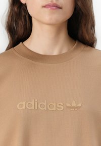 Smešani smeđi dukserica od pamuka s teksturiranim, utisnutim "adidas" logotipom i trefoil dizajnom na prednjoj strani. Riblji izrez s glatkim završetkom.