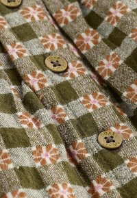 Grøn og beige ternet stof med blomstermønstre. Har brune knapper med indgraveret tekst. Blød tekstur og intrikat vævningsdetalje.