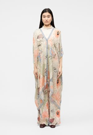 Kvinde iført en løs, flydende blomsterkjole i pastelnuancer med en beige rullekrave under, stående mod en ensfarvet baggrund.