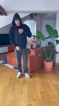 Juodas zip-up džemperis su didele kapišonu, uždėtas ant pilkų kelnių. Modelis stovi ant medinės grindų su vaza ir oranžinės juostelės sofa fone.