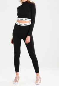 Svart långärmad crop top med vit "Barbie" logotyp på midjebandet, i kombination med svarta leggings och ljusrosa sneakers. Slät, åtsittande design.