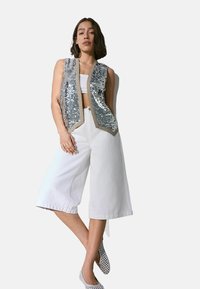 Stříbrná sequined vesta s béžovými lemy, kombinovaná s bílými kalhotami širokého střihu. Obuv je bílá s perforacemi. Jednoduchý, moderní design.
