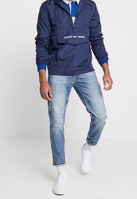 Marinblå vindjacka med fjärdedels dragkedja, framficka och Tommy Jeans-logotyp. Bärs med ljusblå jeans och vita sneakers. Avslappnad stil.