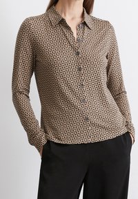 Chemise marron à manches longues avec un motif géométrique beige. Dotée d'un col et de boutons foncés, associée à un pantalon noir.