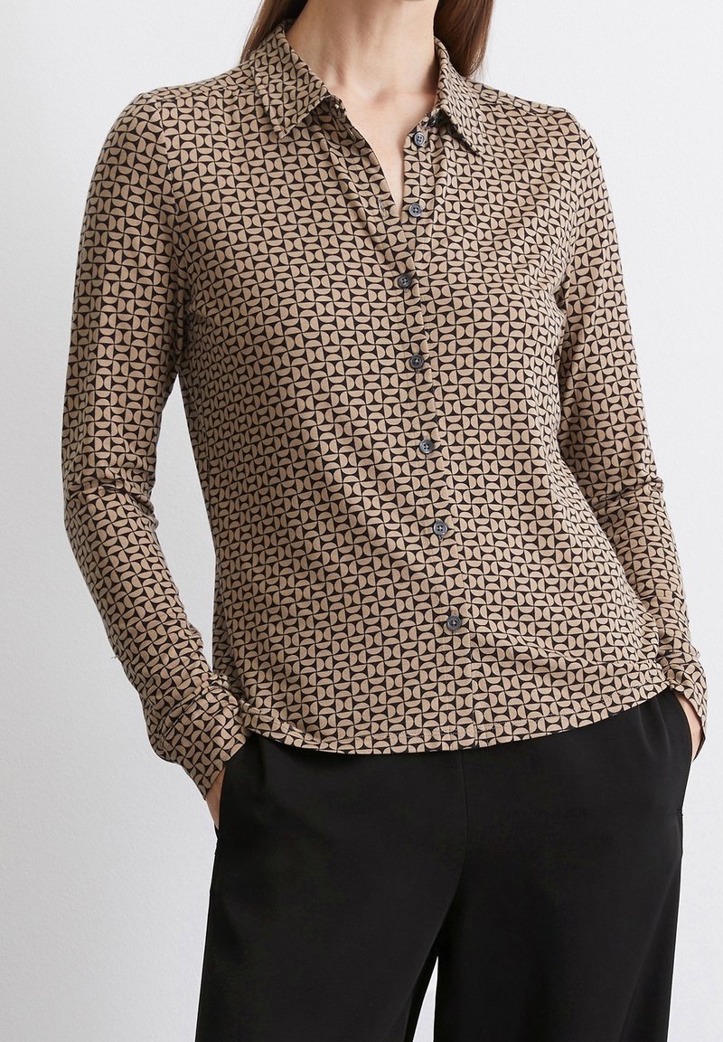 Chemise marron à manches longues avec un motif géométrique beige. Dotée d'un col et de boutons foncés, associée à un pantalon noir.