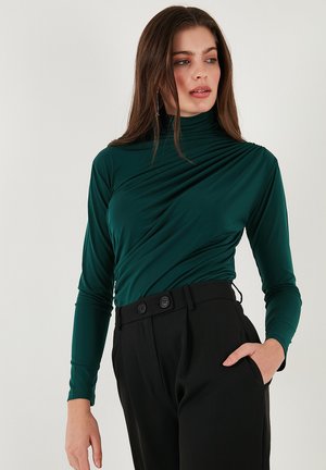Green, long-sleeve top with a high neckline and draped design. Paired with black, high-waisted trousers featuring double-button accents.