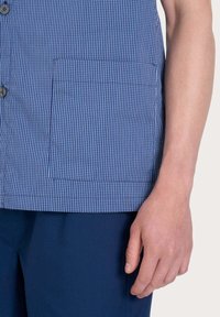 Camicia a scacchi blu e bianca con tasca piatta, chiusura a bottoni e texture liscia, abbinata a pantaloni blu scuro.
