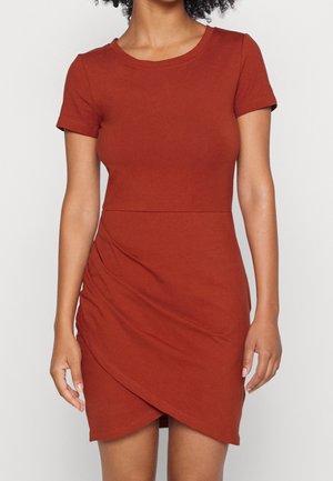 Kobieta ubrana w dopasowaną, rdzawą sukienkę z krótkim rękawem i asymetrycznym efektem kopertowym na spódnicy.