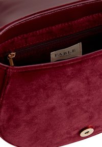 Burgunderfarbene Wildledertasche mit einer weichen Textur. Verfügt über ein Reißverschlussfach und einen Verschluss mit goldfarbener Druckknopf. Im Inneren ist ein Etikett mit der Aufschrift "FABLE" sichtbar.