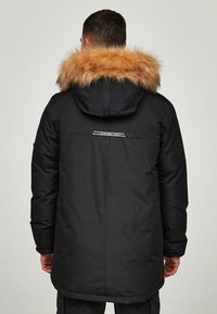 Černá parka s kapucí, která má odnímatelný náplet ze světle hnědé kožešiny. Design zahrnuje hladký povrch a detail loga na zadní straně.