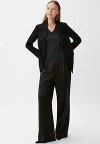 Zwarte op maat gemaakte blazer over een donkergroene V-halstrui, gecombineerd met zwarte wijde broek. Gladde stof met een strakke pasvorm.