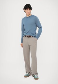 Camisola azul de manga comprida, calças de fato bege claro, cinto castanho com padrão, e ténis verde escuro com detalhes brancos, em pose de pé.