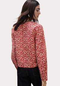Rød mønstret langærmet bluse med hvidt blomstermønster, firkantet udkig, og afslappet pasform, parret med mørk denim. Struktureret stof vist fra ryggen.