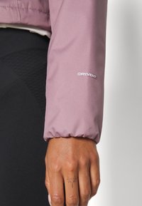 Rosa Dryvent ytterklädesärm med elastisk mud. Närbild av en hand som visar hudtextur och en liten tatuering på fingret.