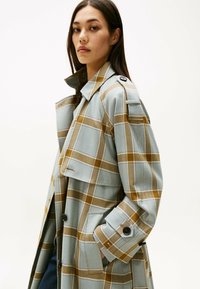 Gabardina con patrón de cuadros en azul claro y marrón, con puños abotonados y bolsillos laterales. La tela parece suave, con diseño clásico de cuello.