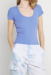 Light purple ribbed shirt with a scoop neckline and short sleeves, paired with white pants featuring a blue tie-dye pattern and pockets.