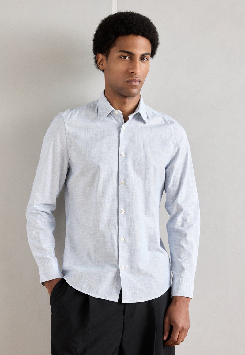 Ανοιχτό μπλε πουκάμισο με κουμπιά και διακριτικές ριγέ υφές, μακρυά μανίκια, μυτερός γιακάς και λευκά κουμπιά, συνδυασμένο με σκουρόχρωμο παντελόνι.