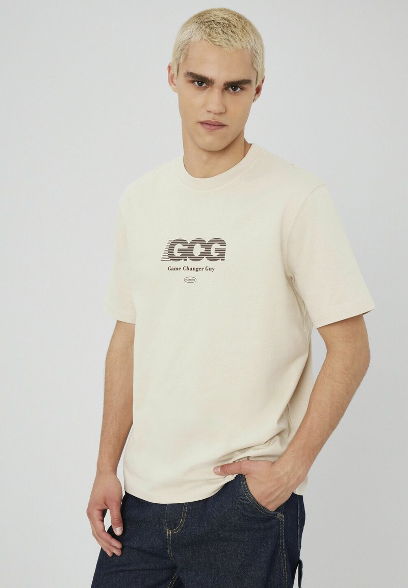 T-shirt en coton couleur crème avec un col rond et des manches courtes, arborant un imprimé graphique en texte noir avec la mention "GCG Game Changer Guy."