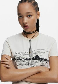 Krémové tričko s čiernou vyšívanou scénou na púšti a textom "Desigual". Má tenký čierny choker so zlatými akcentmi. Hladká textúra.
