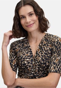 Vzorované šaty s krátkým rukávem, s V-výstřihem a zkrouceným předním dílem. Látka má hnědý a béžový geometrický vzor na tmavém pozadí.