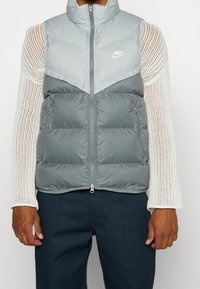 Grå vadderad väst med hög krage, dragkedjestängning och två sidofickor. Har en tydlig färgblockingdesign i över- och underdel. Nike-logotyp på bröstet.