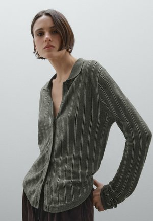 Cardigan à boutons en vert foncé avec des rayures verticales blanches, à manches longues et d'une texture douce. Ses caractéristiques notables incluent une coupe décontractée et un col.