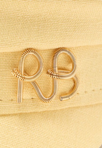 Χρυσό σύρμα σχηματισμένο στα γράμματα "RB", προσαρτημένο σε υφασμάτινη κίτρινη υφή, πιθανώς ως διακοσμητικό λογότυπο ή έμβλημα.