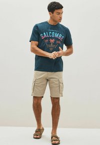 Blue graphic t-shirt with "SALCOMBE" print, cargo shorts in khaki, and brown sandals, displayed against a neutral background.