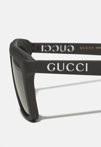 Mustakad prillid paksu raamiga, matt viimistlus ja hallid objektiivid. Külgpinkidel on valgete tähtedega "GUCCI" logo.