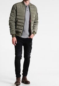 Chaqueta de plumas verde oliva con cierre frontal, puños acanalados y bolsillos laterales, combinada con un polo gris y jeans ajustados negros. Botas marrones hasta el tobillo.