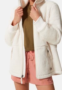 Chaqueta de forro polar blanca con capucha y cremallera frontal, con puños acanalados y bolsillos laterales. Combinada con una camiseta verde olivo y pantalones cortos rosas.