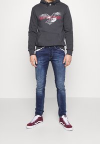 Hombre vestido con sudadera con capucha gris oscuro con gráfico de águila, jeans ajustados azules y zapatillas altas burdeos, de pie contra un fondo blanco.