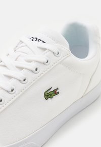 Valkoinen kangaskengät valkoisilla nauhoilla, kumipohjalla ja vihreällä krokodiililogolla sivulla.