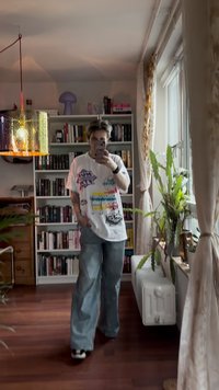 T-shirt gráfica branca oversized com estampados multicoloridos; jeans de perna larga em azul claro; plantas de interior e prateleira ao fundo.