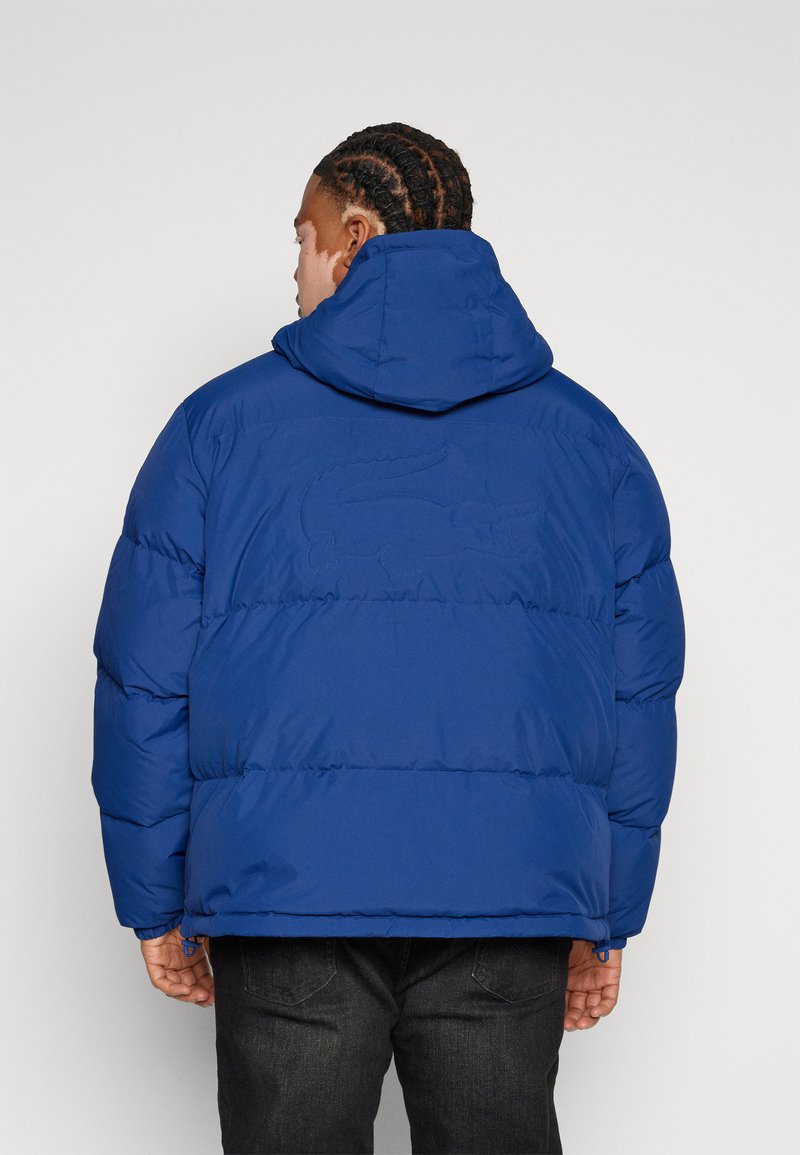 Giacca a vento blu con cappuccio visibile e tessuto strutturato. Presenta un dettaglio del logo cucito sul retro e polsini elasticizzati.