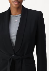 Blazer noir avec col châle, manches longues et ceinture en tissu. Porté par-dessus un top à rayures noir et blanc. Texture lisse, détails minimaux.