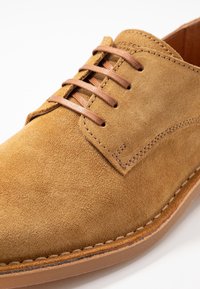 Chaussure en suède beige avec un bout rond, dotée de six œillets en cuir marron pour les lacets et d'une semelle en cuir cousue. Texture lisse et design épuré.