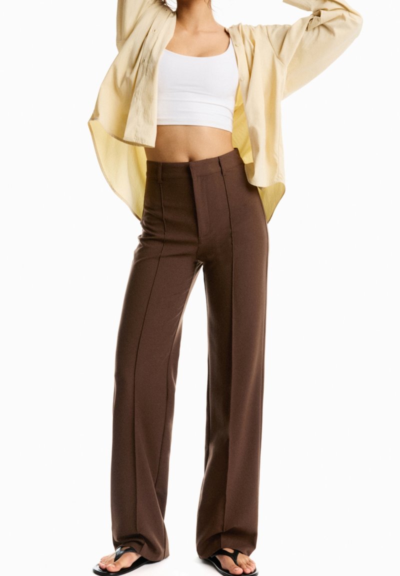 Pantalons marron taille haute avec une coupe ajustée et des coutures verticales, associés à une chemise boutonnée jaune clair par-dessus un crop top blanc.
