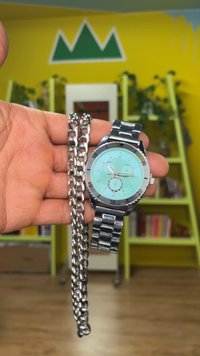 Sidabrinis laikrodis su turkio spalvos ciferblatu, metaline apyranke ir trimis mažais ciferblatais; laikomas šalia sidabrinio grandinėlės apyrankės. Lygus paviršius, elegantiškas dizainas.