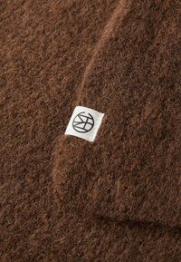Close-up of a brown fuzzy knit fabric with a small white tag featuring a black circular logo with intersecting lines.