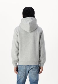 Lys grå hættetrøje lavet af blødt stof, med en stor hætte, ribbede mansjetter og kant. Båret med blå jeans, set fra ryggen.
