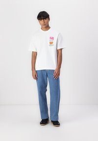 Hvid t-shirt med korte ærmer har grafisk print med "SR" og plante-design. Den er parret med blå lige bukser og sorte sneakers.