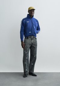 Blå jacka med dragkedja och vita detaljer samt logotyp, grå denimjeans med vita broderade detaljer, svarta skor och en gul keps.