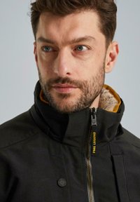 Zwarte jas met een hoge kraag en een zichtbare rits. Gele fleece voering is zichtbaar. Voorzien van een drukknoop en een duurzame stofstructuur.