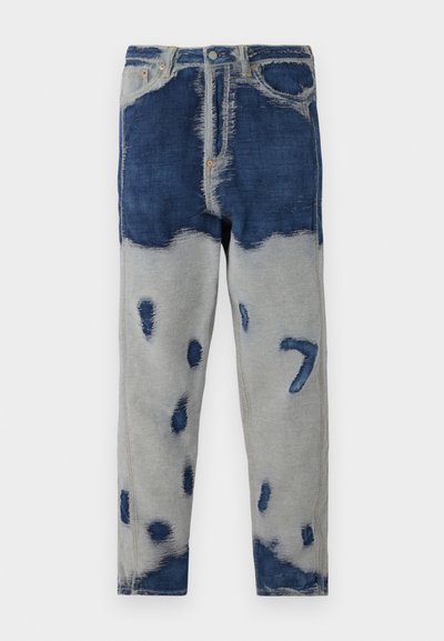 Jean en denim avec un design en patchwork dans des tons de bleu foncé et de gris, présentant des zones usées et des décolorations. Comprend un style classique à cinq poches.